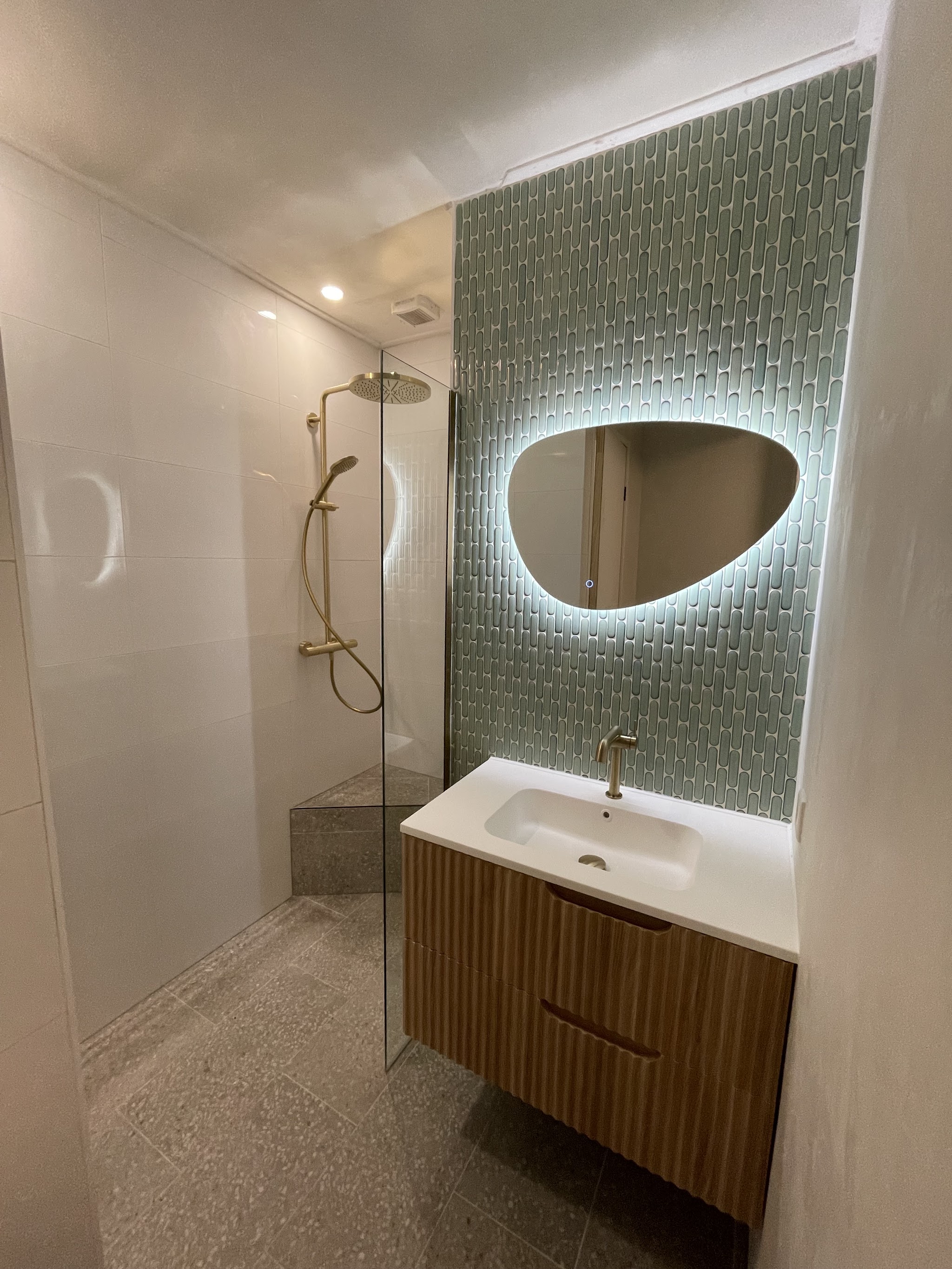Badkamer met groene reliëftegels, verlichte spiegel, houten badmeubel en gouden regendouche